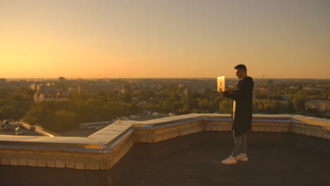 middle plan: a male freelancer typing on a laptop keyboard at sunset overlooking the city while standing on the roof of a skyscraper. the businessman works remotely and controls the work of the company