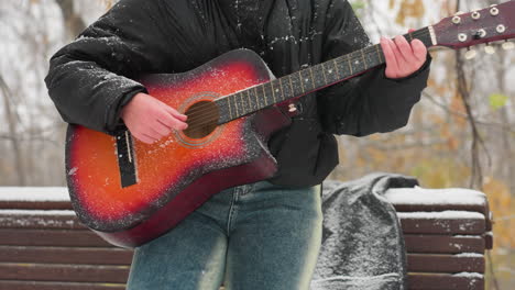 primer plano de manos tocando una vibrante guitarra acústica roja y naranja al aire libre en un parque nevado, la nieve cubre las cuerdas y la manga, mezclando la belleza serena del invierno con la pasión musical