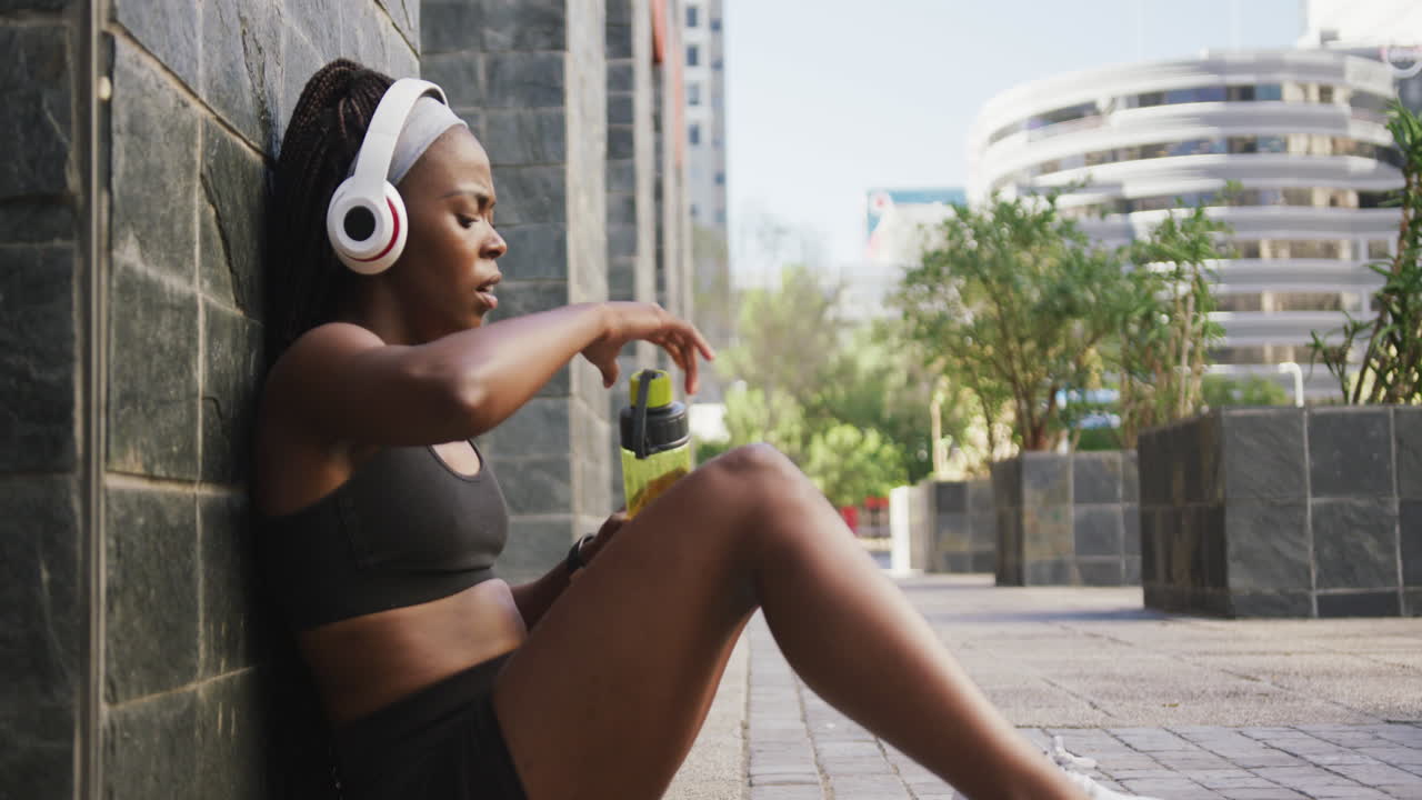 African american woman exercising outdoors wearing wireless headphone drinking water in the city