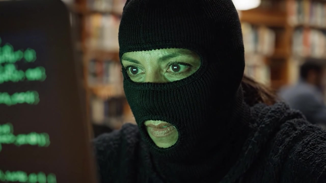 En mystisk maskeret skikkelse udfører spændende hackeraktivitet i et bibliotek, omgivet af bøger og digitale koder, der lyser på laptopskærmen.