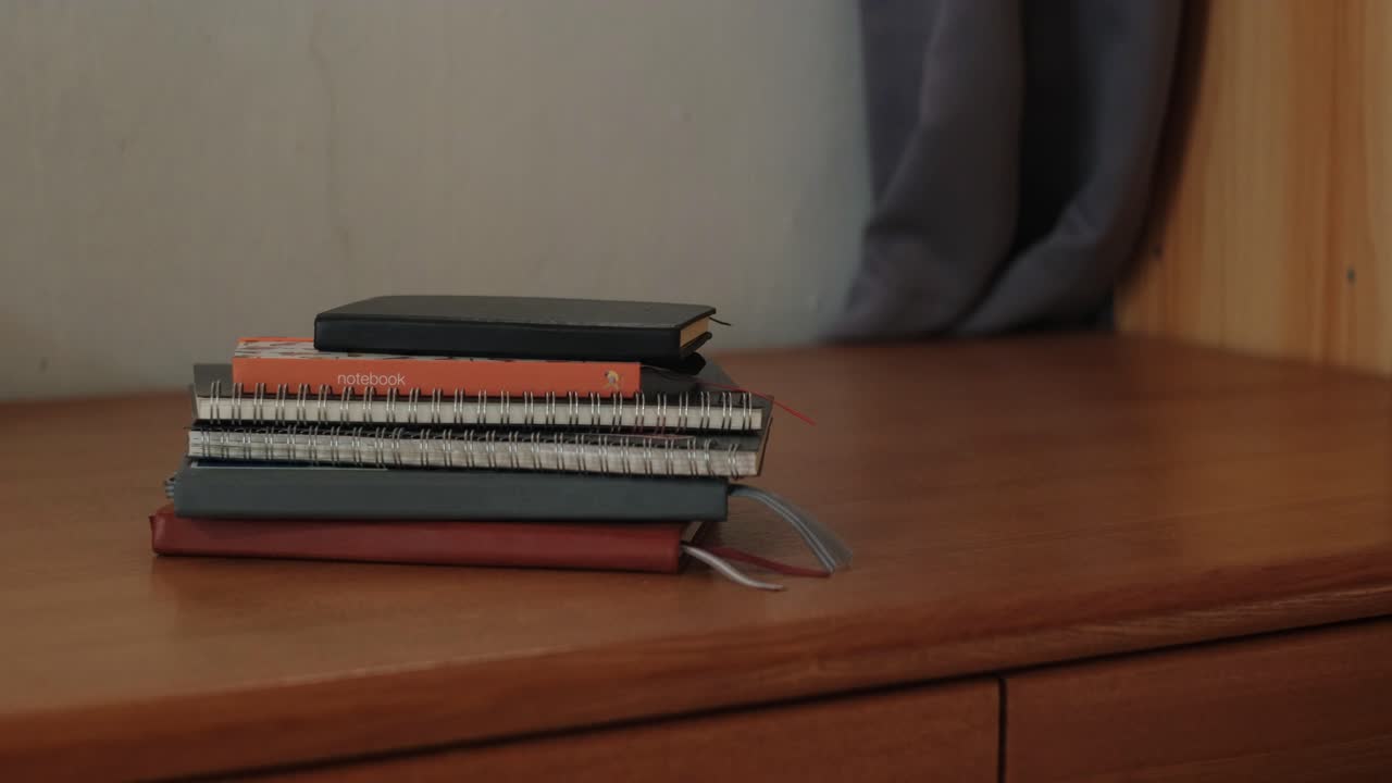 Static medium shot of pile of notebook journals atop wooden desk as hand grabs one from top