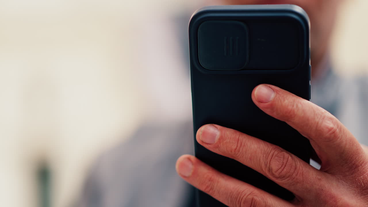 Close up of a man's hand holding a smartphone with a black case and a camera cover on a blurred background