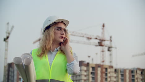 Sprechende-Frau-Mit-Helm-Am-Telefon-Vor-Dem-Hintergrund-Einer-Baustelle-Mit-Kränen,-Die-Zeichnungen-In-Der-Hand-Halten.-Ingenieurin-Auf-Der-Baustelle