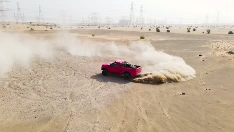 Vista-De-Drones-En-Cámara-Lenta-De-Un-Camión-Rojo-Girando-Neumáticos-Haciendo-Un-Donut-Levantando-Una-Nube-De-Polvo-En-Una-Zona-Desértica-Arenosa