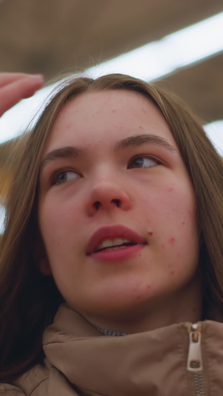 Close-up shot of a young girl wearing a brown jacket, gently touching her hair while in a mall. The background is blurred, emphasizing the girl's thoughtful expression and casual gesture