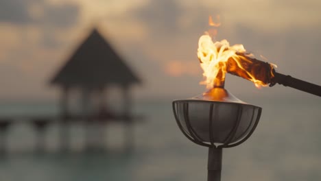 Lámpara-De-Aceite-De-Acero-En-El-Poste-Iluminada-Por-Una-Antorcha-Encendida-Con-Una-Espectacular-Puesta-De-Sol-En-El-Fondo