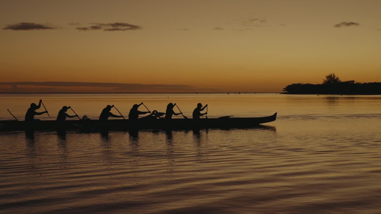 a 6 person team paddles an outrigger canoe or wa'a across a calm ocean in the Hawaiian islands at sunset as other boats follow behind silhouette