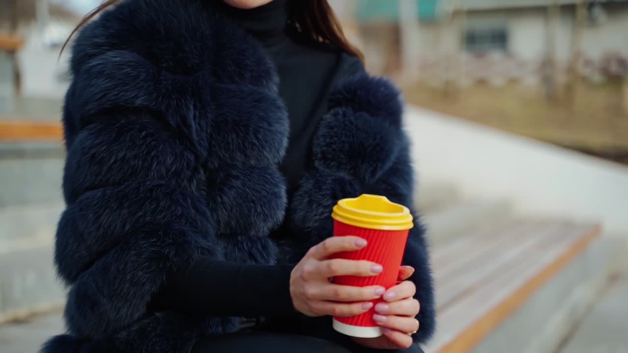Female hands hold a cup of beverage outdoors. Beautiful woman in a fur coat sitting in the park and drinking from plastic glass. Slow motion.