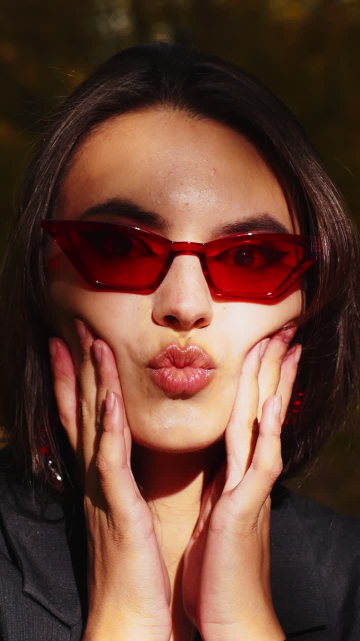 Stylish Model Posing with Red Sunglasses, Showcasing Bold Facial Expressions Against a Vibrant Background During an Outdoor Photoshoot