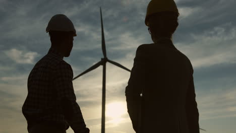 Silhouetten-Einer-Kaukasischen-Frau-Und-Eines-Ingenieurs,-Die-Einen-Helm-Tragen,-Die-Hände-Schütteln-Und-In-Die-Kamera-Einer-Windstation-Für-Erneuerbare-Energien-Blicken