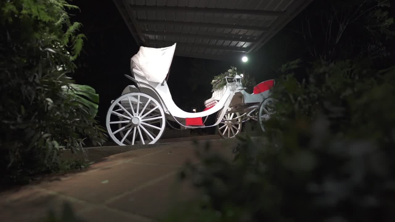 A wide shot of an elegant white carriage prop decorated with flowers and lit by soft event lighting. The scene captures the romantic ambiance of an outdoor wedding at night