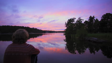 Rising-crane-shot-over-a-man-sitting-at-a-rocky-shore,-of-a-lake,-a-purple-sky,-at-a-colorful-sunset-or-dusk,-at-Albysjon,-Tyreso,-Sweden