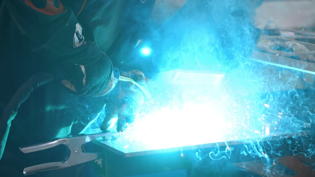 Worker in welder's mask welding metal parts with blue high-temperature sparkles - slow motion