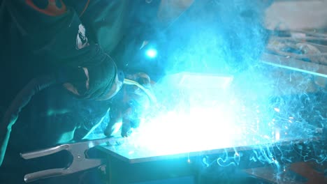 worker in welder's mask welding metal parts with blue high-temperature sparkles - slow motion