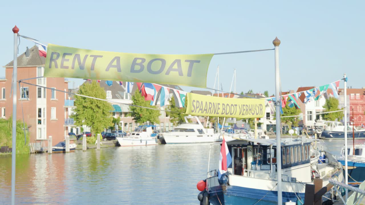 A bright daytime view of a canal in Haarlem, Netherlands, with a 'Rent a Boat' banner above moored boats and Dutch architecture. Static camera, natural lighting
