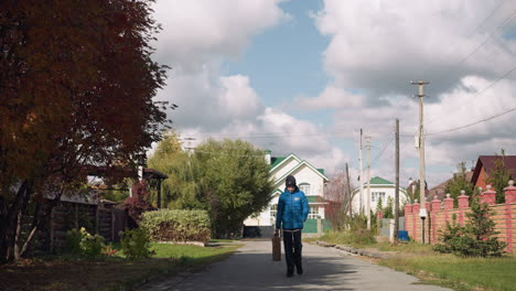 teenager in blue hoodie and black beanie carries paper bag, strolling down suburban street lined with autumn foliage and distinctive homes under a cloudy sky