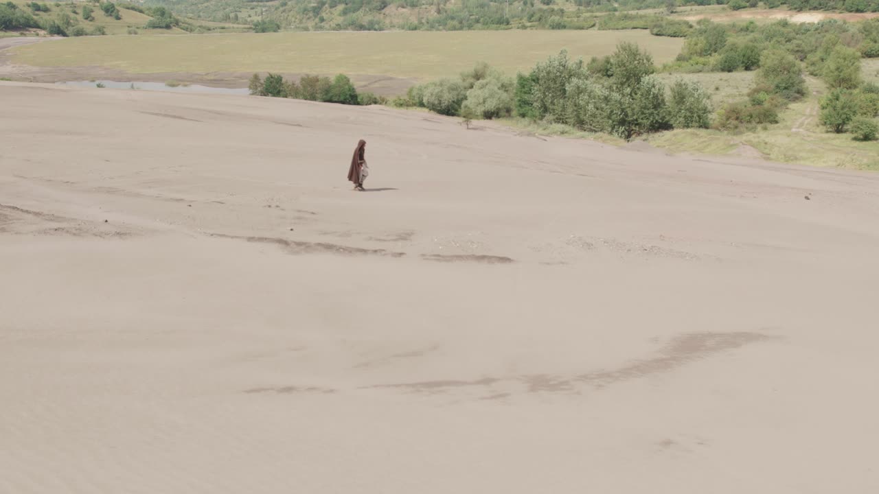 A solitary robed figure walks across a barren, post-apocalyptic wasteland. In the background, a lush green valley offers a stark contrast. A concept of a long journey or survival