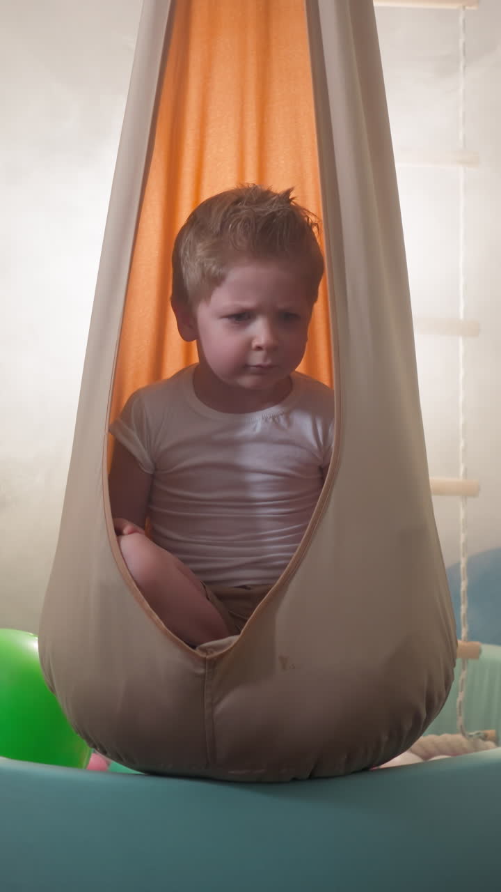 Cute blond toddler frowns moving head to sides in apartment gym. Little boy with suspicious facial expression sits in hanging hammock in room slow motion