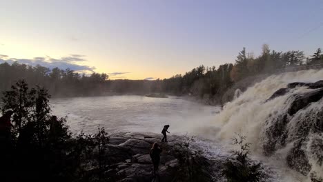 Menschen,-Die-Bei-Sonnenuntergang-Im-Wald-Auf-Rutschigen-Felsen-Neben-Einem-Riesigen-Und-Heftigen-Wasserfall-Laufen