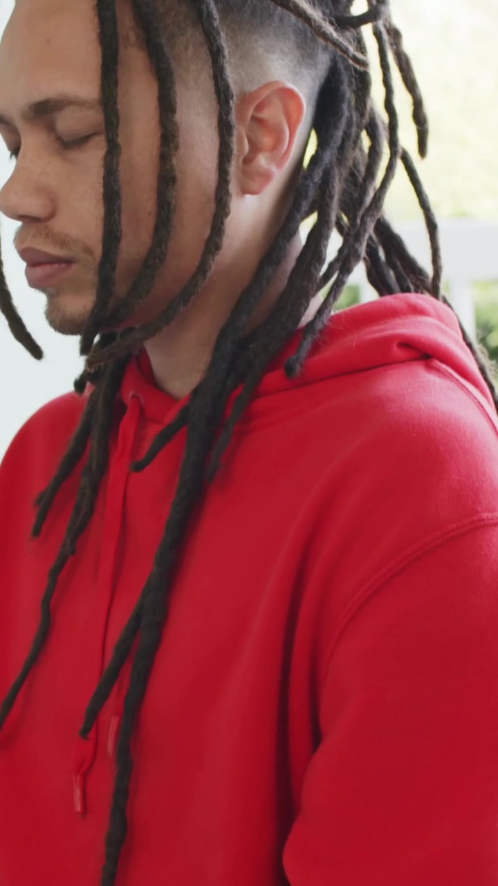 Video verticale: Telecamera panoramica che mostra un uomo seduto che si calma in veranda, con felpa rossa e dreadlocks.