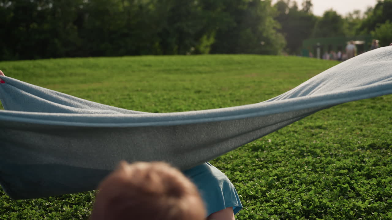 youngsters playing around picnic blanket on grass while adults and families stroll near playground, summer evening light, laughter and movement in park, joyful children sharing playful moment