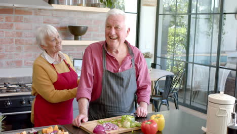 happy senior caucasian couple wearing aprons cooking dinner in kitchen at home, slow motion