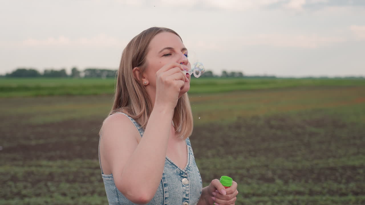 young woman in sleeveless denim dress blows soap bubbles from wand while standing on dusty rural path beside wide green field under soft evening sky, droplets glinting around swirling