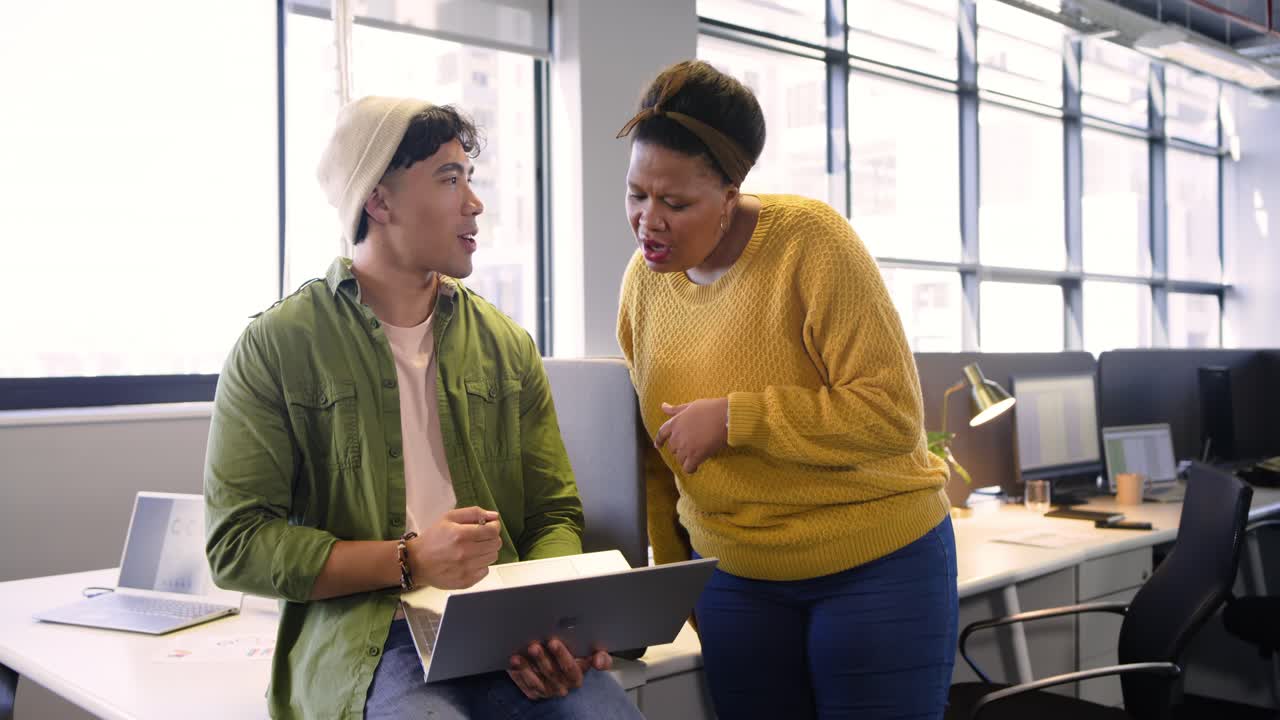 Diverse coworkers in office, woman walking in and pointing at laptop while man reviewing design