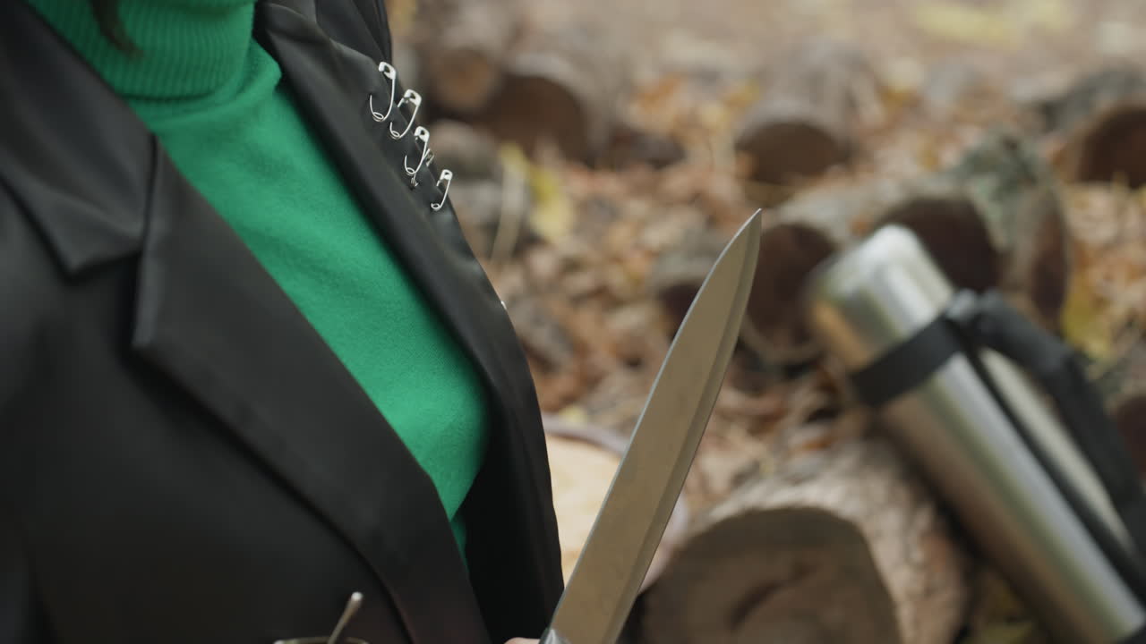 Woman leather jacket holding knife outdoors, green sweater visible, hands securing buckle and sliding blade from sheath near stacked logs and autumn leaves thermos on log, closeup of blade and grip