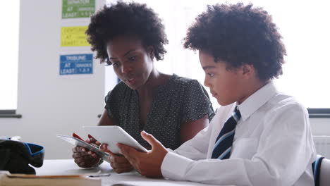 female high school tutor with digital tablet giving male student wearing uniform one to one tuition at desk