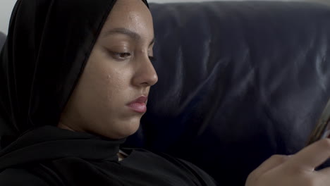 close up side view of young muslim women wearing hijab typing on smartphone whilst sat on sofa