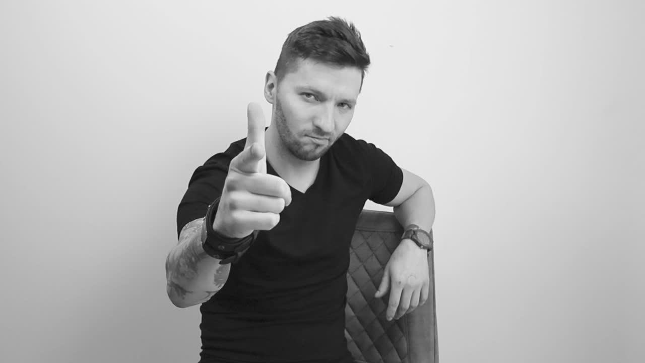 Filmato in scala di grigi di un uomo tatuato, con capelli corti e barba, seduto su una sedia, che guarda direttamente la telecamera, alza il dito puntando in avanti e annuisce con approvazione e sorride