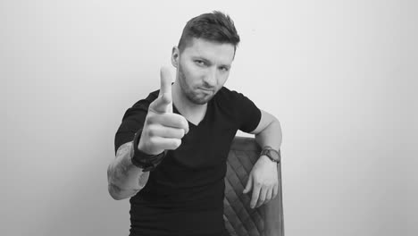 grayscale footage of a tattooed short haired and bearded man sitting on a chair, looking directly forward at the camera and lifting his finger pointing forward and nodding with approval and smiling.