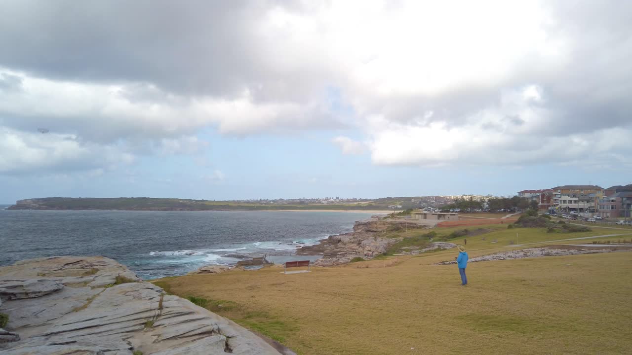 파란색 옷을 입은 남자가 바람이 많이 부는 흐린 날 해변 근처에서 모형 원격 제어 항공기를 가지고 놀고 있습니다.