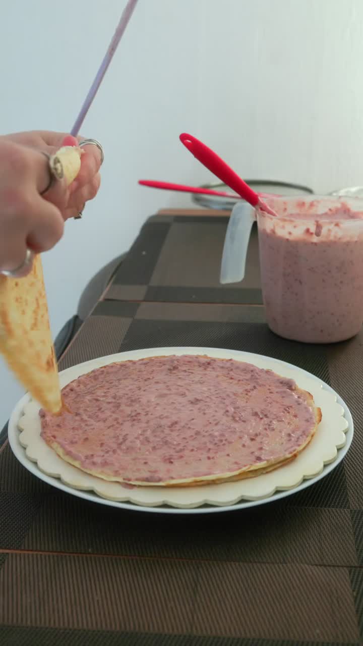 Woman's hands layering a crepe cake with raspberry cream on white plate. Close up vertical side view