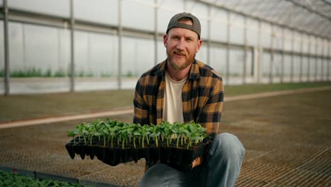 Portrait-of-a-happy-guy-farmer-in-a-cap-in-a-plaid-shirt-who-holds-seedlings-of-young-plants-in-his-hands-in-a-greenhouse-on-the-farm