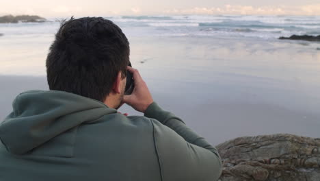 young man in green hoodie in left of frame seated quietly on seaside rocks at the beach during early morning, staring at the calm ocean waves while drinking a coffee from an insulated travel mug