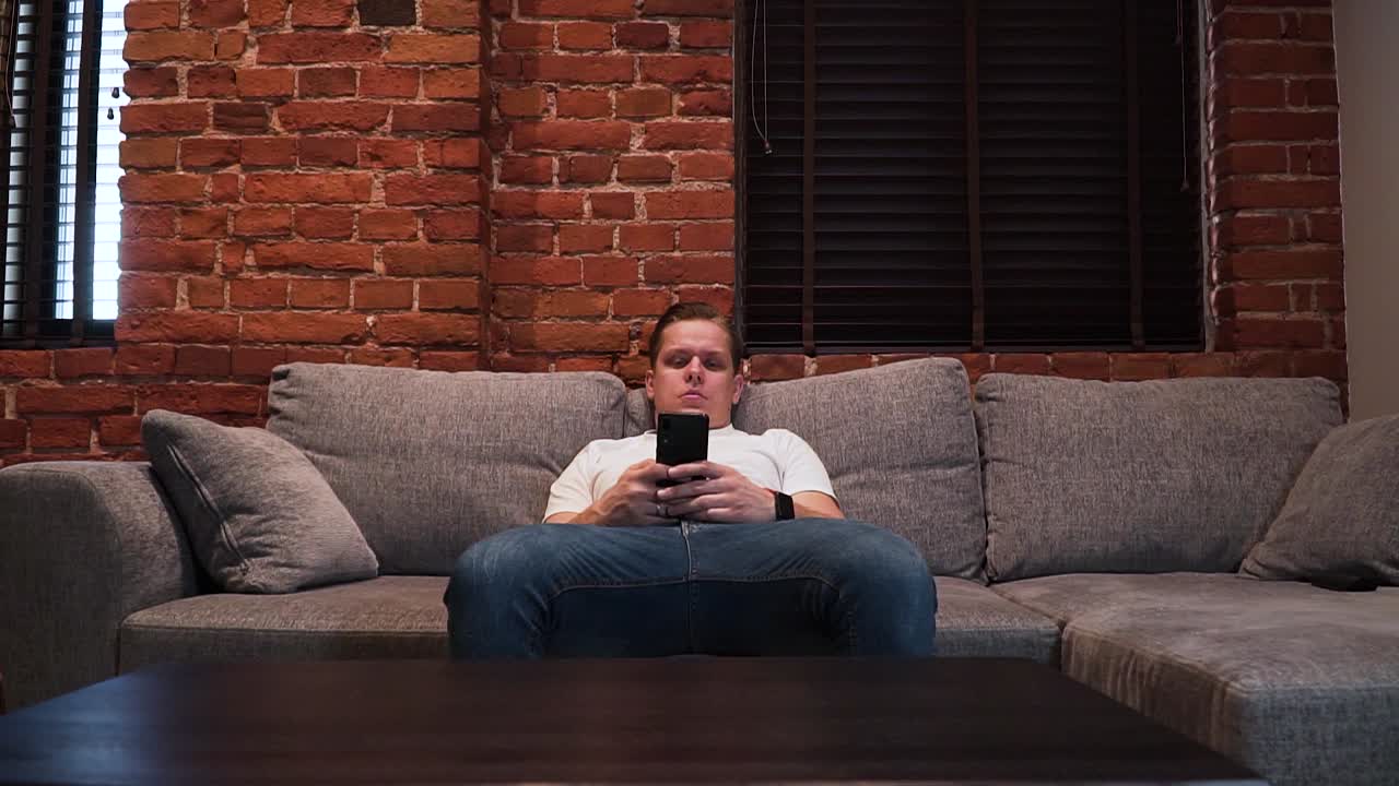 진지해 보이는 젊은이가 거실 소파에 앉아 정면에서 찍은 스마트폰으로 메시지를 쓰고 있다