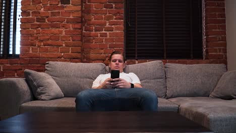 the young serious-looking man is sitting on the couch in the living room and is writing a message with his smartphone, shot from the front