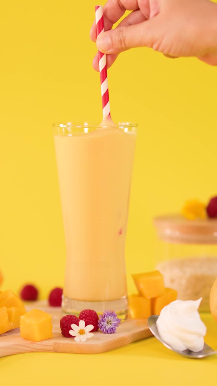 Una mano mescola un frullato alto al mango con una cannuccia a righe, circondato da frutta fresca su uno sfondo giallo vivace.