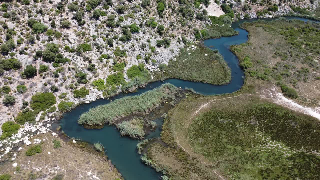 Aerial drone shot showing meandering stream winding through rocky and marshy terrain near Koycegiz Wetlands in Mugla with green patches, textured landscape, calm water bends, and bright daylight