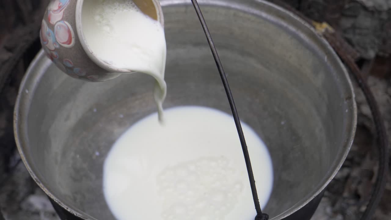 Cup of milk poured into a black cauldron and set on fire. The cup is made of clay with different colors. The cauldron is above the fire made in nature among the woods