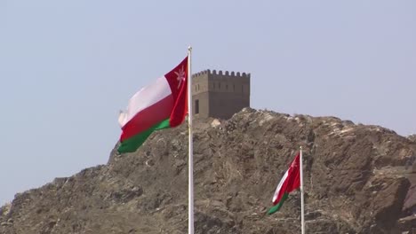 two flags of the state of oman waving in the wind in the back you can see the tower of the fortress