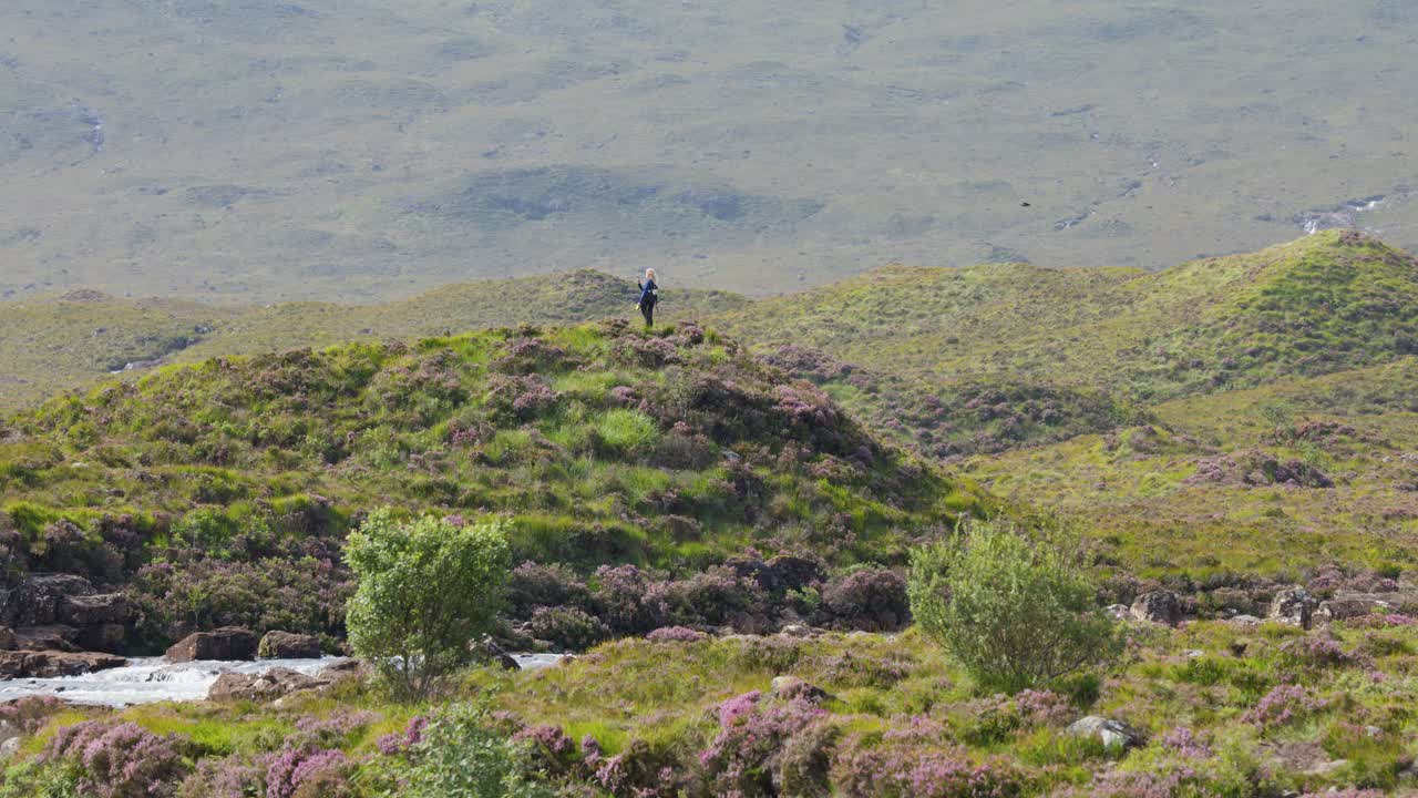 Single hiker pauses atop green hill, wide landscape view, natural daylight, steady camera, tranquil mood