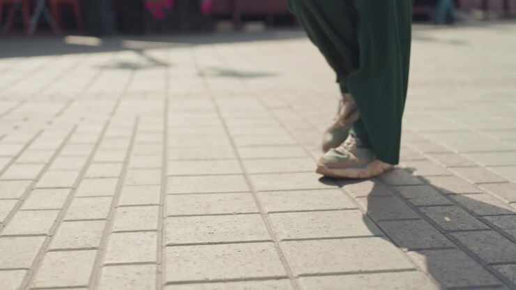close up feet crossing sunlit plaza, green trousers and beige sneakers casting long shadow, blurred crowd background, confident pace, rhythmic stepping, textured paving, candid city capture, everyday