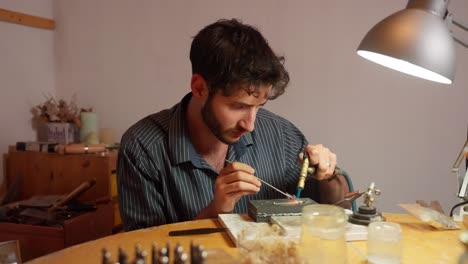 un artesano se centra en detallar una pieza de joyería en una mesa de madera. la luz cálida ilumina las herramientas y materiales esparcidos por ahí.