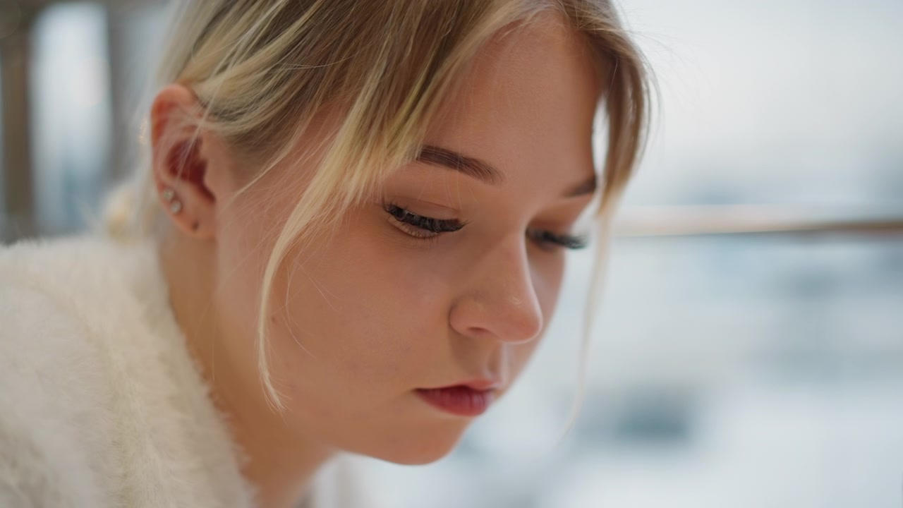 Close-up head and shoulders view of young woman tilting head downward with lowered gaze and relaxed lips, while soft daylight from window and slight hair movement create calm contemplative mood