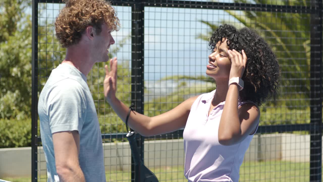 Diverse couple on padel court flirting, man offering paddle strap, woman taking and adjusting watch