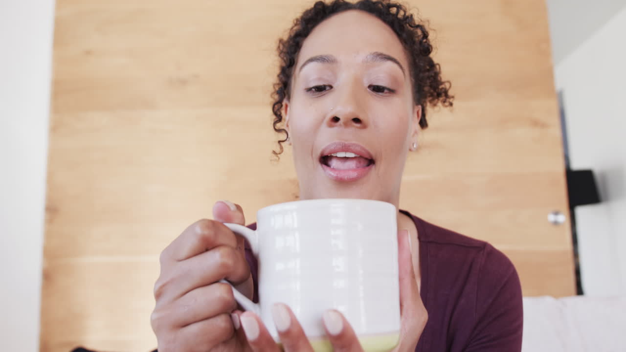 mujer biracial feliz haciendo una llamada de video hablando y bebiendo café en la sala de estar, en cámara lenta