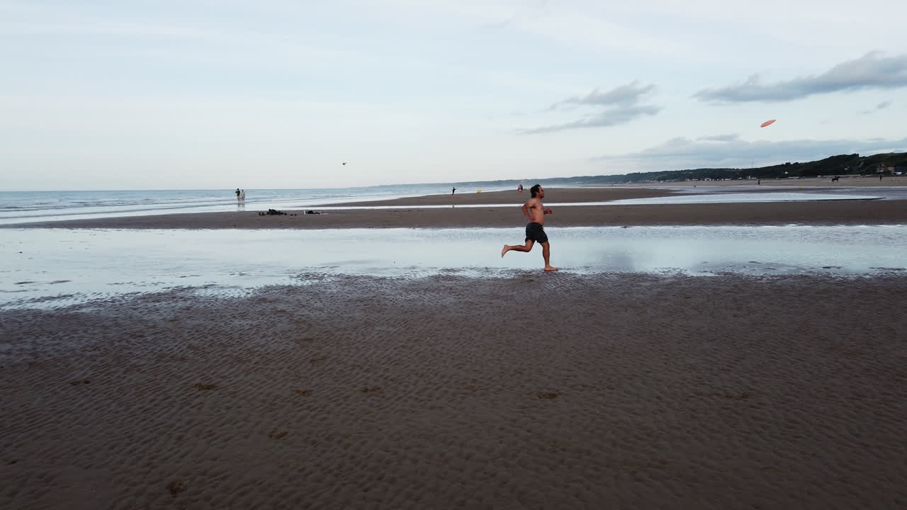 Shirtless man throws frisbee on Omaha Beach, France. Casual beach game on a historic shoreline, blending playful energy with a peaceful, expansive coastal landscape.